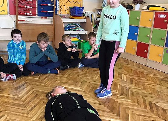 Dziewczynka podchodzi do poszkodowanej dziewczynki i wykonuje kolejne czynności. Reszta dzieci obserwuje z zainteresowaniem.