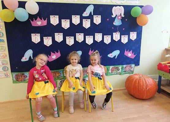 Trzy dziewczynki siedzą na krzesełkach na tle gazetki ściennej