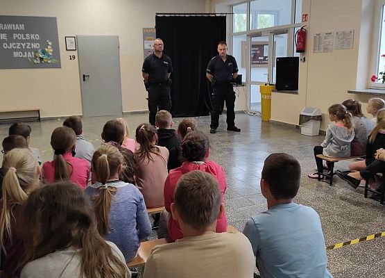 Uczniowie siedzą na ławkach i słuchają policjantów