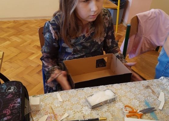 Uczennica siedzi przy stoliku i trzyma na kolanach karton