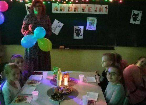 Dzieci siedzą przy stoliku a obok stoi nauczycielka z balonami
