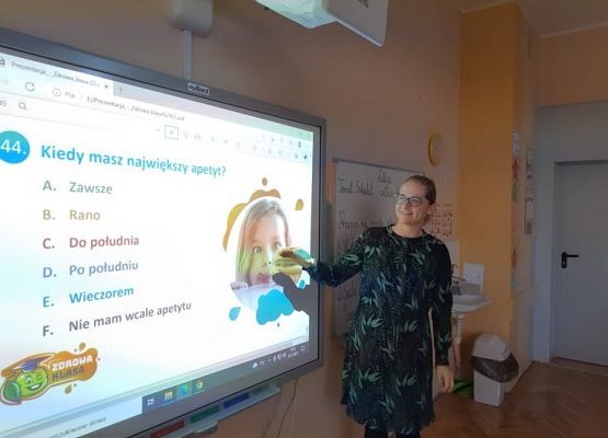 Nauczycielka stoi przy tablicy i odczytuje dzieciom ankietę