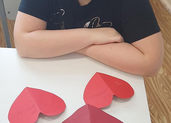 Uczennica siedzi w ławce, na blacie leżą trzy czerwone, papierowe serca