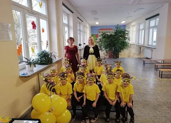 Grupa przedszkolaków przebranych za pszczółki siedzi na ławkach. Za dziećmi stoją dwie panie.