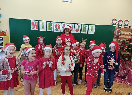 Grupa dzieci i pani nauczycielka stoją na tle tablicy. Wszyscy w  mikołajkowych przebraniach