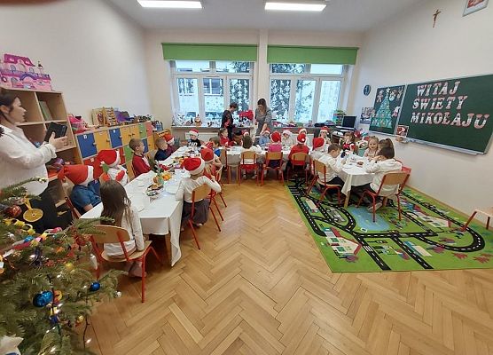 Dzieci siedzą przy stole, nauczycielka rozdaje dzieciom opłatek