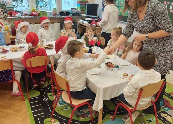 Dzieci siedzą przy stole, nauczycielka rozdaje dzieciom opłatek