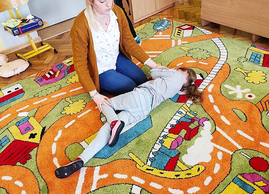 Dziewczynka leży na podłodze a nauczycielka pokazuje dzieciom, jak należy ułożyć dziewczynkę w bezpiecznej pozycji