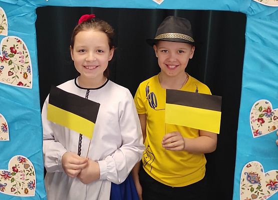 Uczniowie stoją w ramce, na której są wzory kaszubskie. Trzymają flagi kaszubskie.