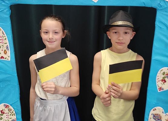 Uczniowie stoją w ramce, na której są wzory kaszubskie. Trzymają flagi kaszubskie.