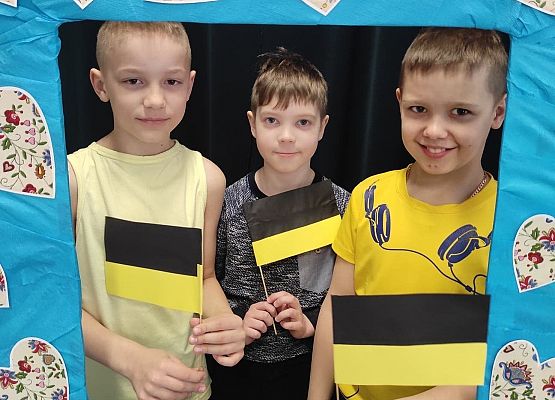 Uczniowie stoją w ramce, na której są wzory kaszubskie. Trzymają flagi kaszubskie.