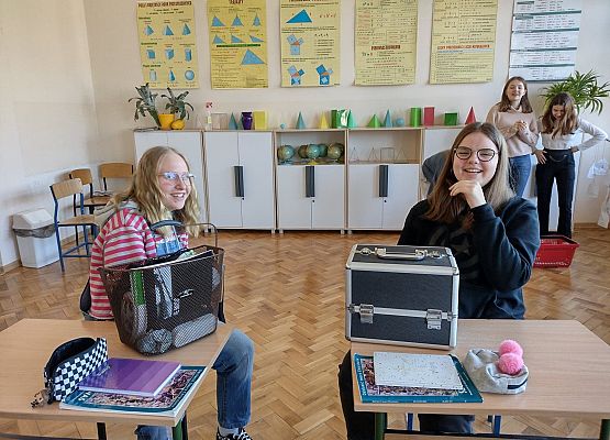 Uczennice siedzą przy ławce pokazując, w czym przyniosły książki