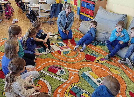 Uczniowie siedzą na dywanie i słuchają nauczycielki