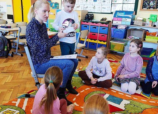 Uczniowie siedzą na dywanie i słuchają nauczycielki