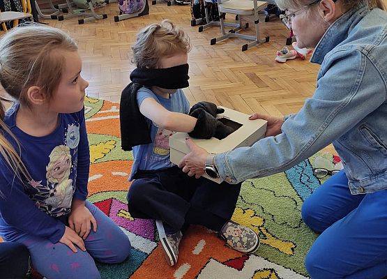 Nauczycielka podaje  przedmioty do rozpoznania dziewczynce z zasłoniętymi oczami