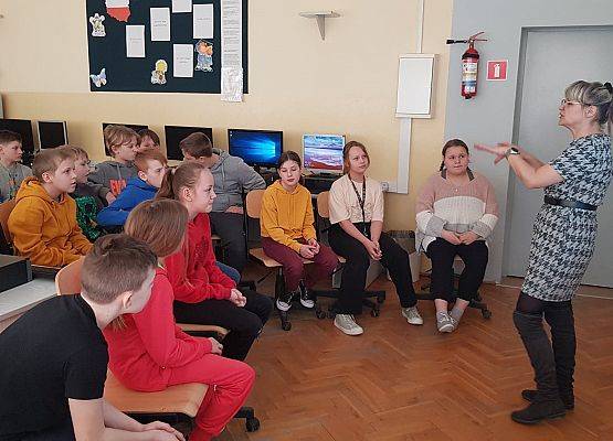 Uczniowie siedzą na krzesełkach a nauczycielka stoi i opowiada