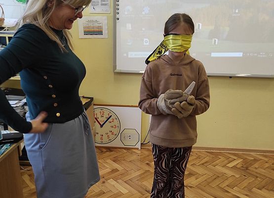 Uczennica z zakrytymi oczami na na dłoniach robocze rękawice, obok stoi nauczycielka