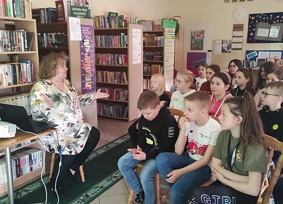 Uczniowie siedzą w bibliotece i słuchają nauczycielki