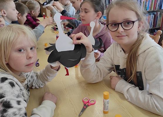 Dzieci siedzą przy stole i wykonują papierowe bociany