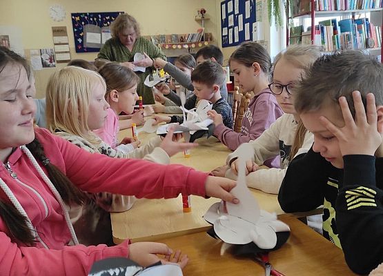 Dzieci siedzą przy stole i wykonują papierowe bociany