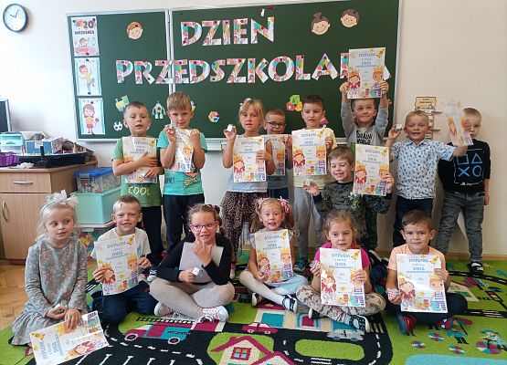 Dzieci stoją przy tablicy z napisem Dzień Przedszkolaka i pokazują dyplomy