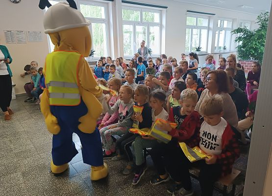 Dzieci siedzą na ławkach, obok stoi Mrówka
