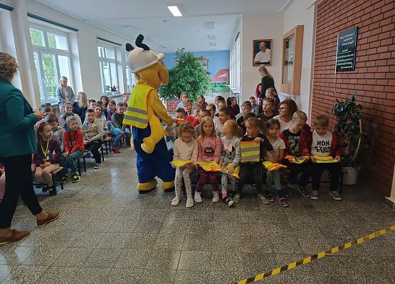 Dzieci siedzą na ławkach, obok stoi Mrówka