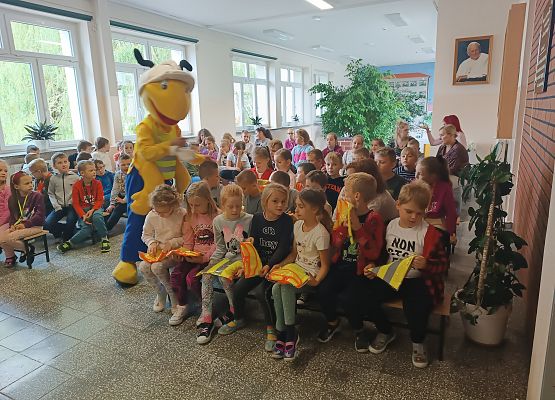 Dzieci siedzą na ławkach, obok stoi Mrówka