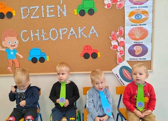 Chłopcy siedzą na krzesełkach na tle tablicy z napisem dzień chłopaka