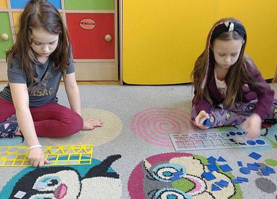dwie dziewczynki siedzą na dywanie i dopasowują figury matematyczne