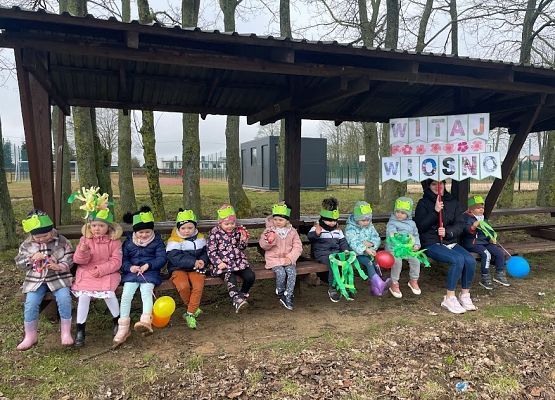 dzieci odpoczywają na ławce, pani trzyma transparent z napisem witaj wiosno