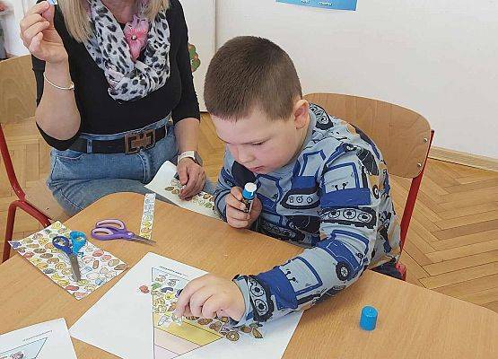 chłopiec przykleja obrazki na kartkę, obok siedzi nauczycielka
